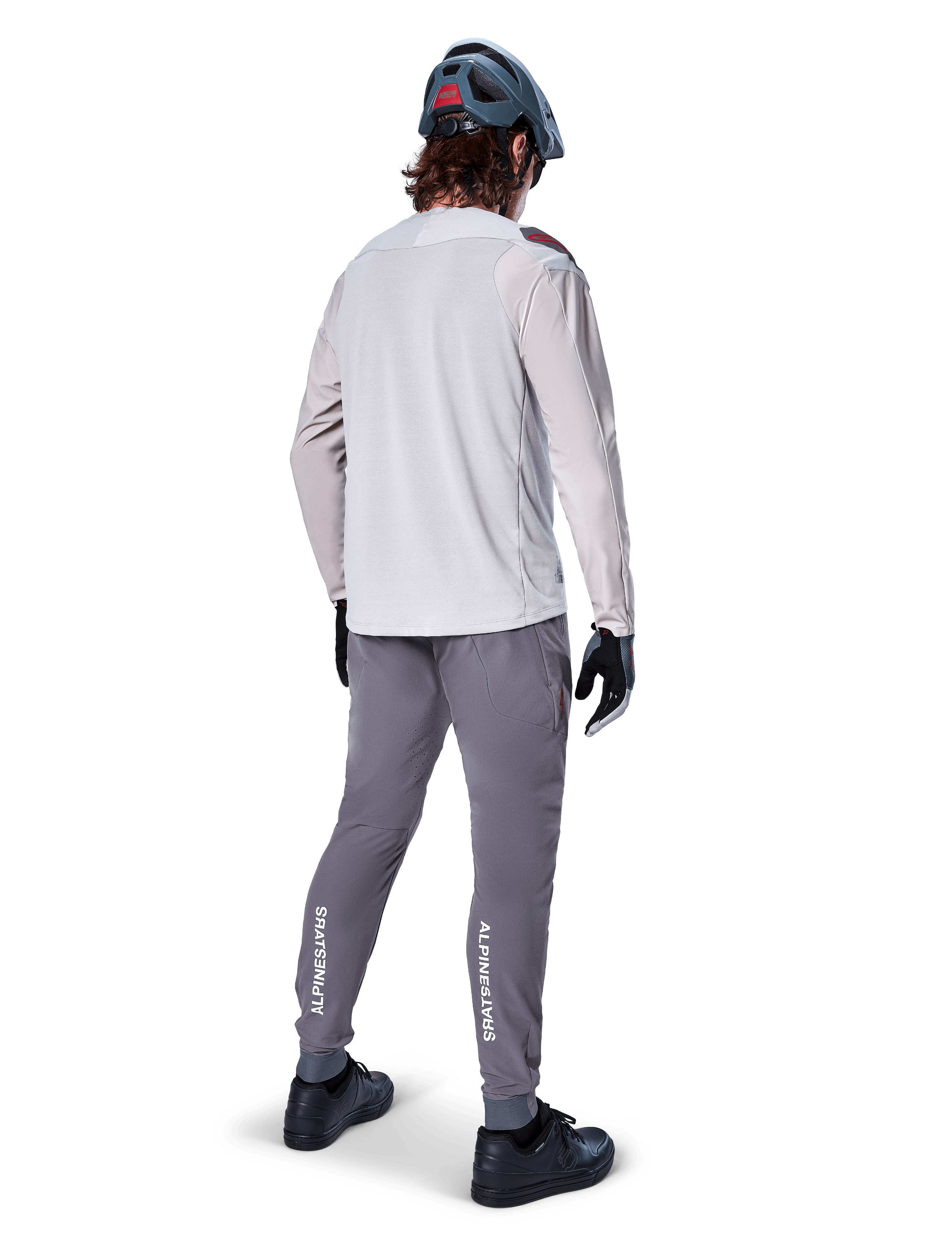 Vista posteriore di un ciclista che indossa la maglia a maniche lunghe Alpinestars A-Aria Polartec Switch in grigio chiaro, abbinata a pantaloni da mountain bike grigio scuro con marchio Alpinestars bianco sui polpacci, guanti neri e un casco grigio