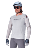 Alpinestars A-Aria Polartec Switch Jersey a maniche lunghe, maglia da ciclismo grigio chiaro con logo nero sul petto e dettaglio rosso sulla spalla, maniche perforate al laser per la ventilazione, indossata con casco azzurro, occhiali da sole e pantaloni grigi, abbigliamento tecnico da mountain bike per trail
