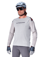 Alpinestars A-Aria Polartec Switch Jersey a maniche lunghe, maglia da ciclismo grigio chiaro con logo nero sul petto e dettaglio rosso sulla spalla, maniche perforate al laser per la ventilazione, indossata con casco azzurro, occhiali da sole e pantaloni grigi, abbigliamento tecnico da mountain bike per trail