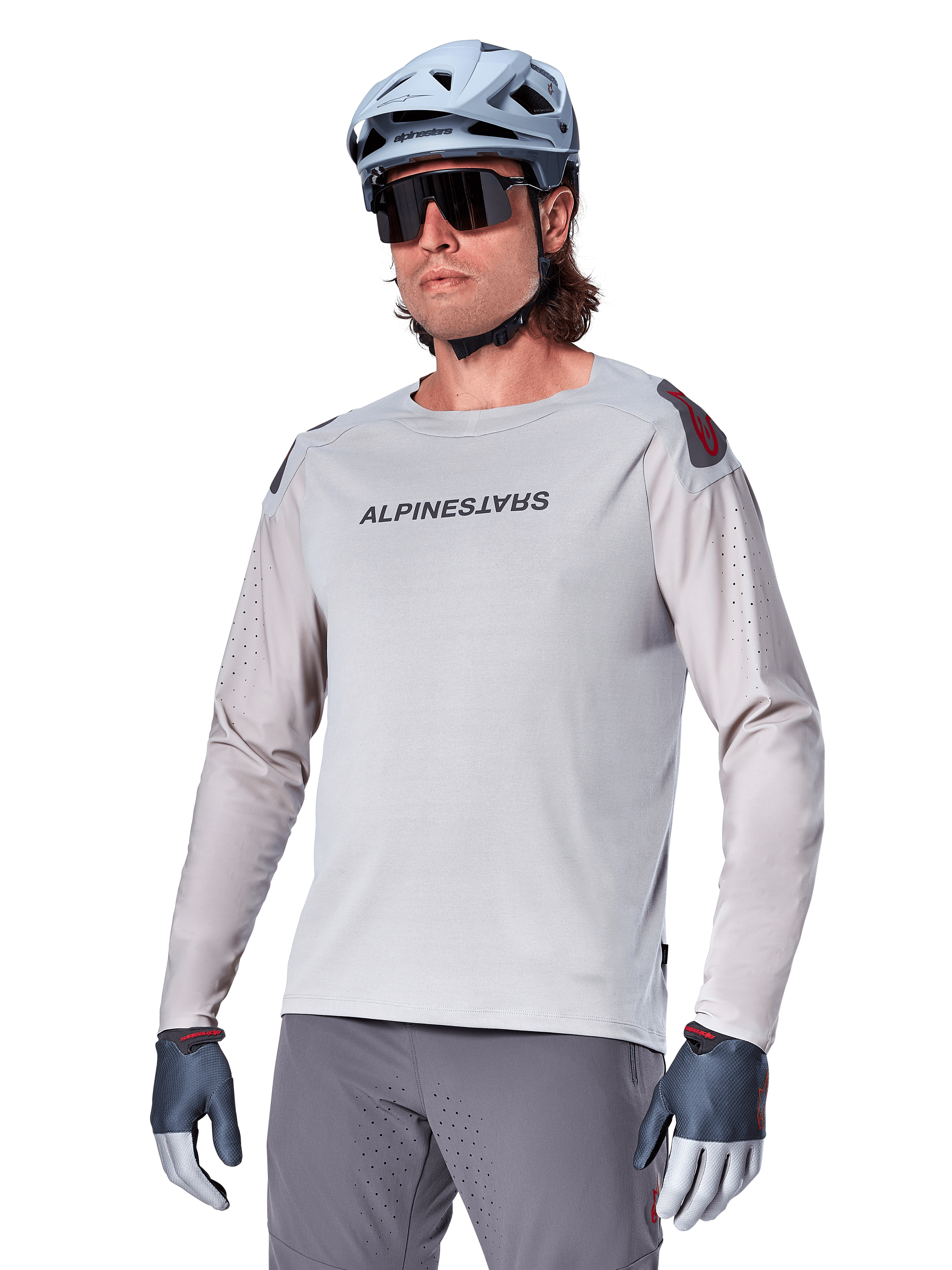 Alpinestars A-Aria Polartec Switch Jersey a maniche lunghe, maglia da ciclismo grigio chiaro con logo nero sul petto e dettaglio rosso sulla spalla, maniche perforate al laser per la ventilazione, indossata con casco azzurro, occhiali da sole e pantaloni grigi, abbigliamento tecnico da mountain bike per trail