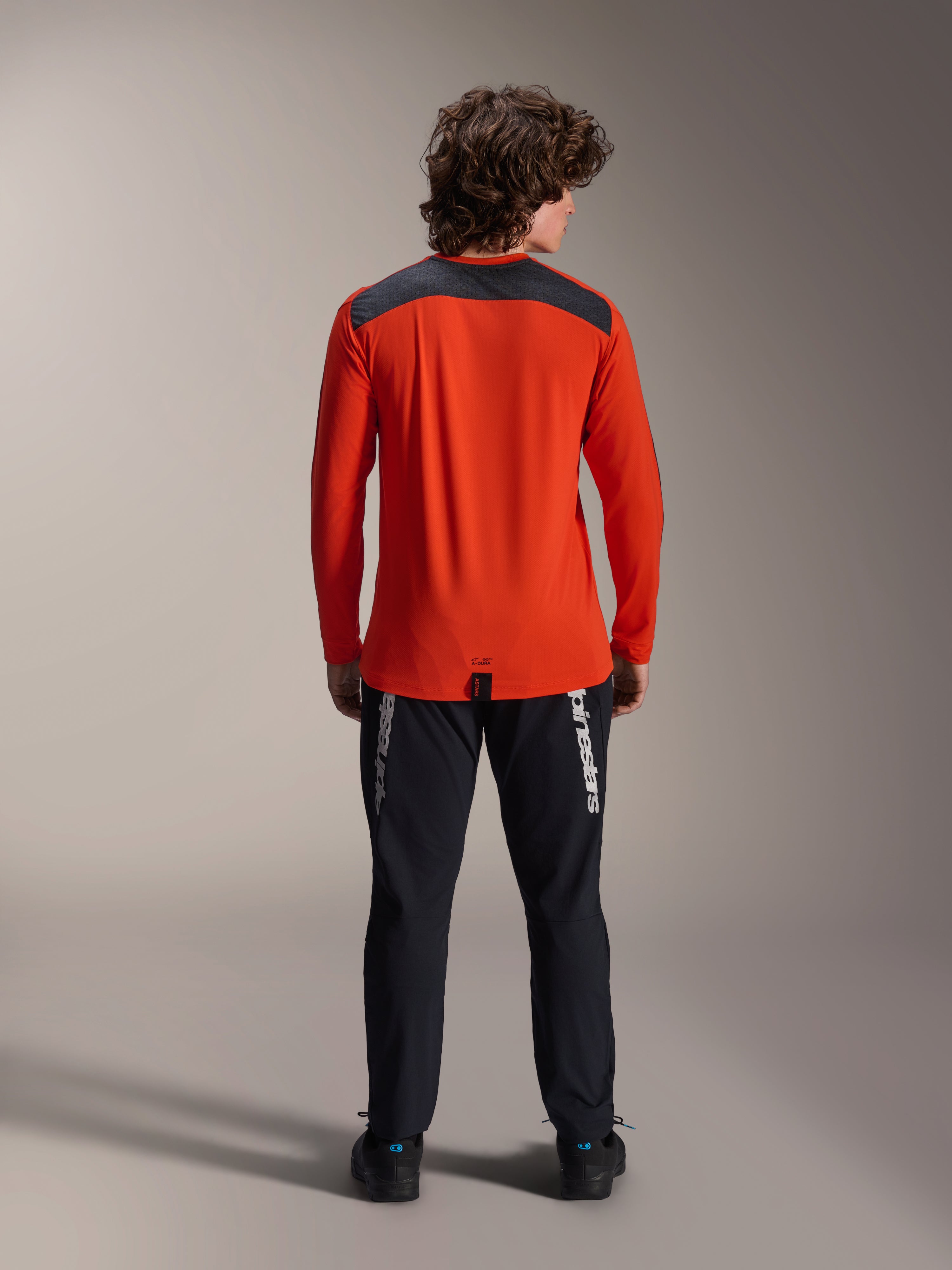 Maglia Alpinestars A-Dura Insert Astar a maniche lunghe, Spicy Orange, vista posteriore con pannello sulle spalle grigio antracite, indossata con pantaloni Alpinestars neri e scarpe nere, scatto professionale in studio per abbigliamento tecnico da MTB