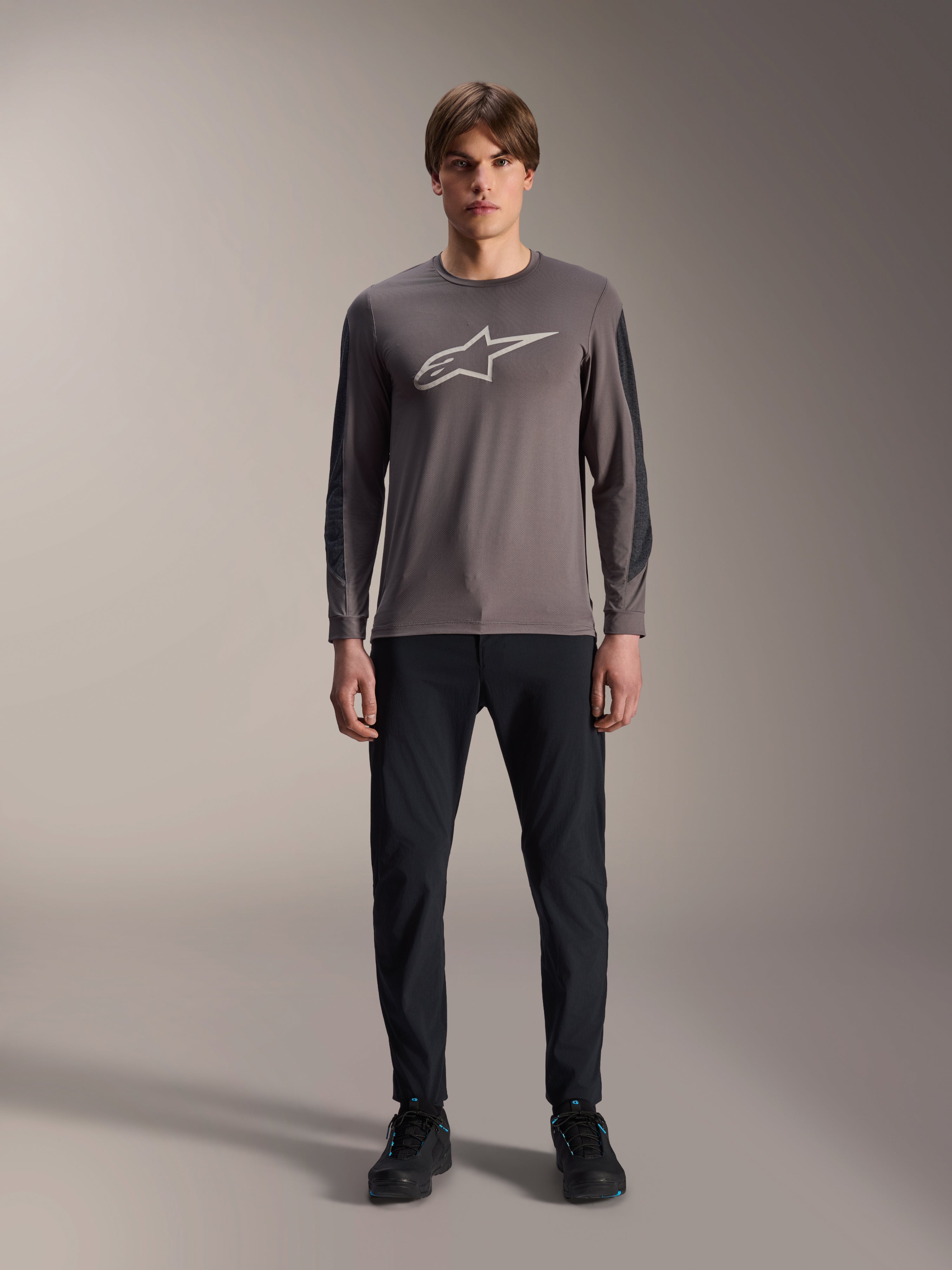 Alpinestars Maglia A-Dura Insert Astar, maglia da MTB a maniche lunghe, grigio scuro, con logo Astar grigio chiaro sul petto e inserti testurizzati più scuri sulle maniche, indossata da un modello con pantaloni e scarpe neri, abbigliamento tecnico per mountain bike