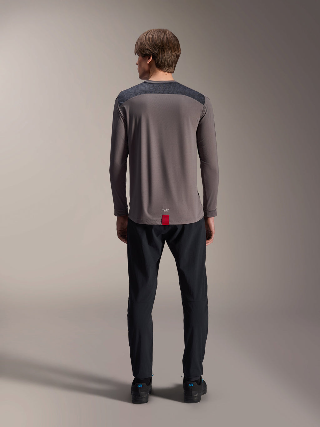 Maglia a maniche lunghe Alpinestars A-Dura Insert Astar in grigio scuro, vista posteriore, con pannelli sulle spalle color antracite e un'etichetta rossa sull'orlo, indossata con pantaloni e scarpe neri, abbigliamento tecnico per mountain bike