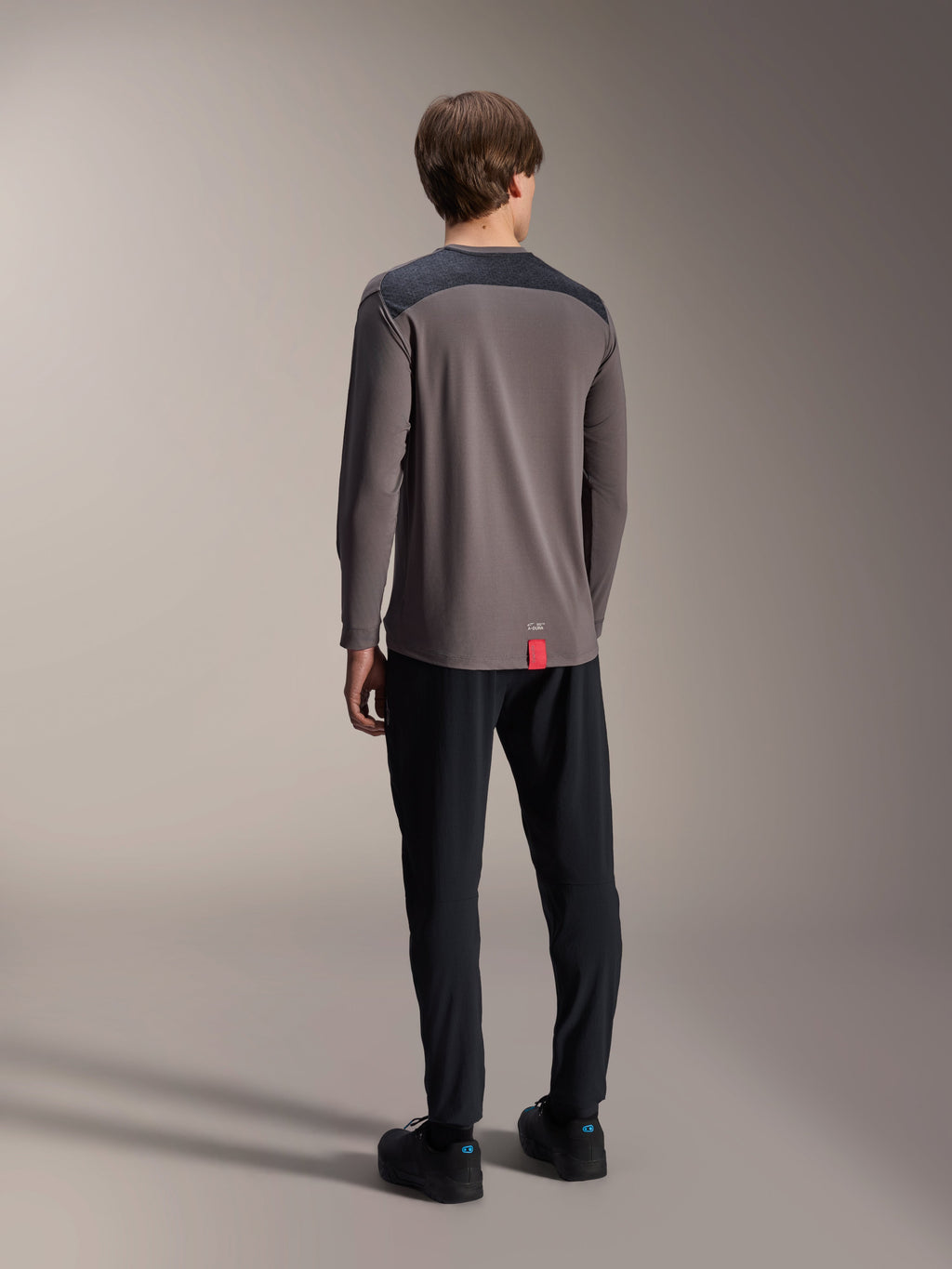 Maglia a maniche lunghe Alpinestars A-Dura Insert Astar, grigio scuro, vista posteriore su modello, con pannelli rinforzati sulle spalle color antracite e una piccola etichetta rossa sull'orlo, abbinata a pantaloni e scarpe neri, abbigliamento tecnico da MTB