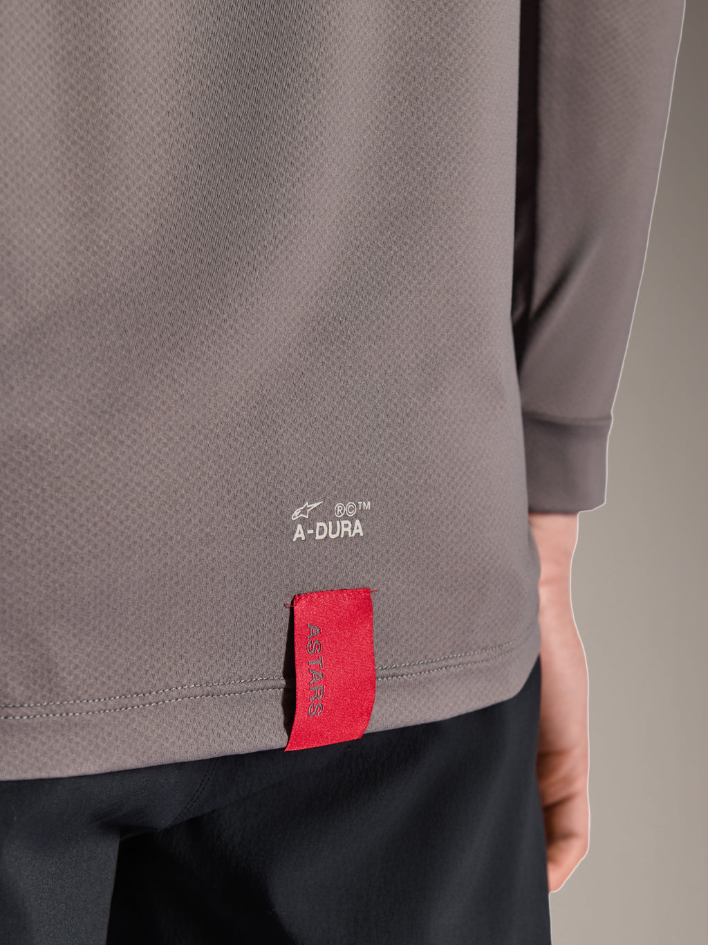 Alpinestars A-Dura Insert Astar Jersey, maglia MTB a maniche lunghe, grigio scuro, dettaglio ravvicinato dell'orlo posteriore con marchio A-DURA bianco ed etichetta in tessuto rossa ASTARS, tessuto tecnico testurizzato