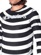 Primo piano della maglia Alpinestars A-Aria Oscar a maniche lunghe in nero, con strisce bianche orizzontali, grafica a spirale Oscar e logo Alpinestars argento riflettente sul petto, indossata da un pilota con cinturino del casco visibile.