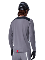 Alpinestars Maglia A-Dura Dri Astar a maniche lunghe, maglia da MTB grigio scuro vista dal retro, con pannello nero sulle spalle e piccola etichetta rossa del brand, indossata con casco e guanti da mountain bike neri