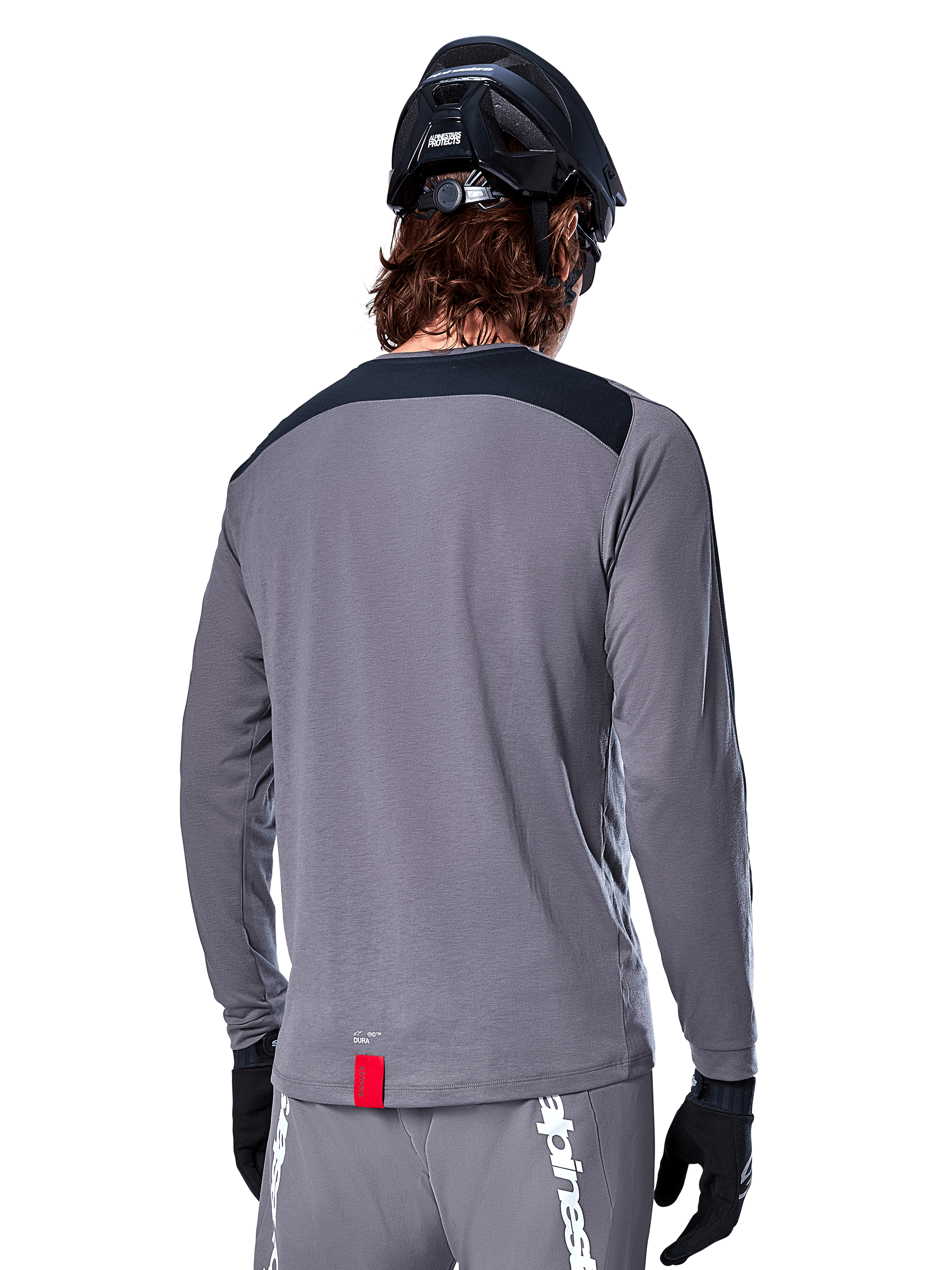 Alpinestars Maglia A-Dura Dri Astar a maniche lunghe, maglia da MTB grigio scuro vista dal retro, con pannello nero sulle spalle e piccola etichetta rossa del brand, indossata con casco e guanti da mountain bike neri