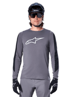 Alpinestars A-Dura Dri Astar Long Sleeve Jersey, maglia a maniche lunghe grigio scuro, con un grande logo Astar bianco sul petto e pannelli in mesh neri sulle braccia, indossata da un ciclista con casco da mountain bike nero e occhiali tecnici, abbigliamento da ciclismo MTB