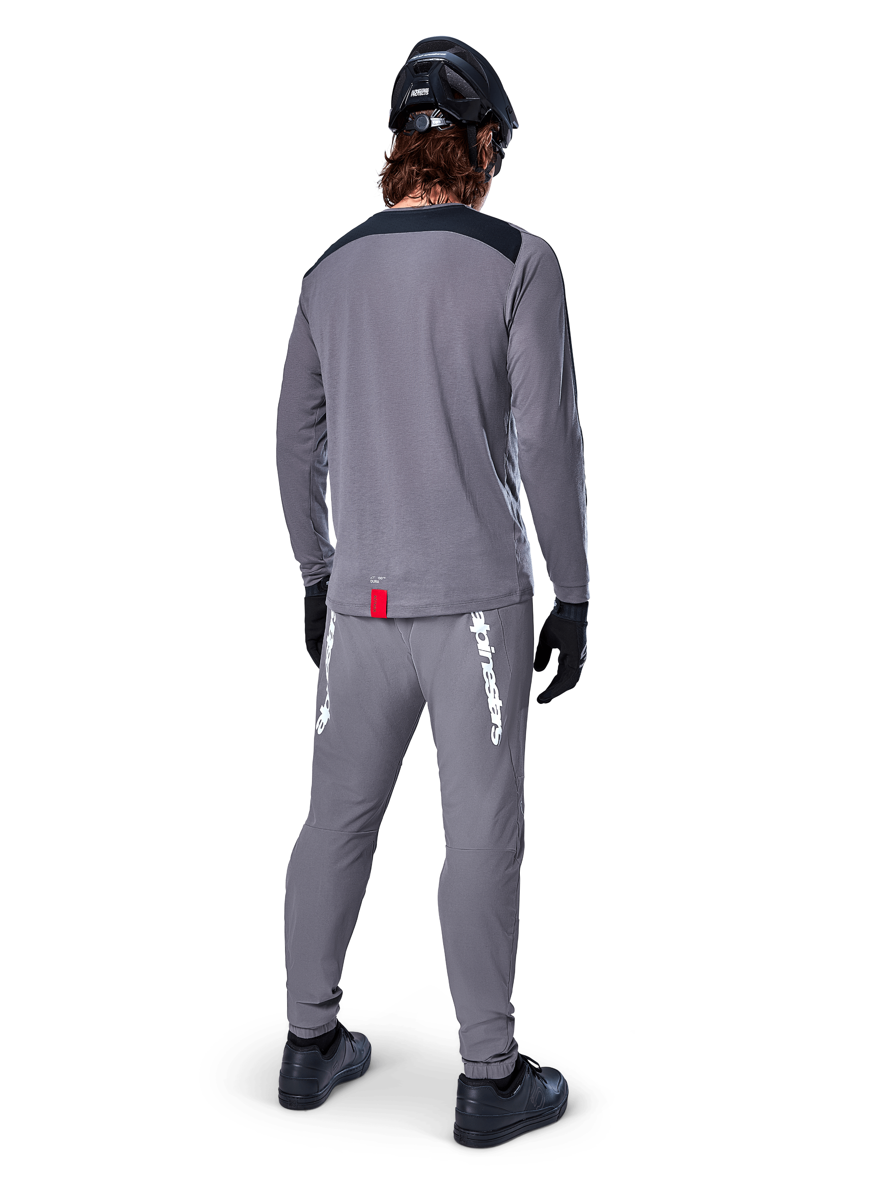 Maglia Alpinestars A-Dura Dri Astar - Maniche Lunghe, grigio scuro, vista posteriore del ciclista con pantaloni abbinati e casco nero, presenta pannelli neri sulle spalle e una piccola etichetta rossa sull'orlo, abbigliamento tecnico da ciclismo fuoristrada