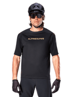 Mountain biker con maglia a maniche corte Alpinestars A-Aria Polartec Switch in nero, logo dorato sul petto, abbinata a casco da MTB nero, occhiali da sole tecnici scuri e guanti coordinati, abbigliamento da ciclismo fuoristrada.