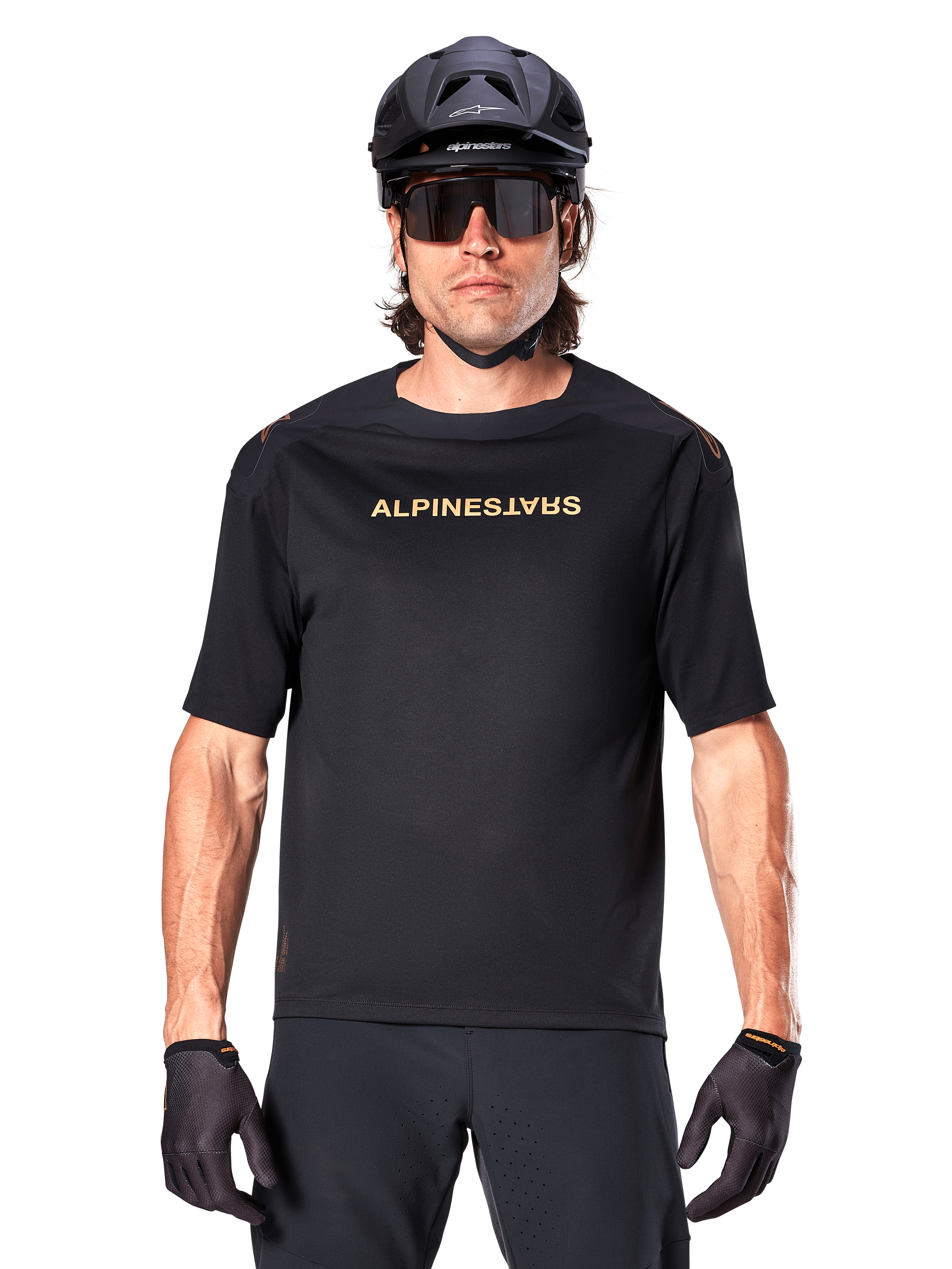 Mountain biker con maglia a maniche corte Alpinestars A-Aria Polartec Switch in nero, logo dorato sul petto, abbinata a casco da MTB nero, occhiali da sole tecnici scuri e guanti coordinati, abbigliamento da ciclismo fuoristrada.