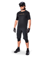 Alpinestars A-Aria Polartec Switch Short Sleeve Jersey, maglia a maniche corte, nero con marchio dorato specchiato sul petto, indossata con pantaloncini da ciclismo neri, casco e ginocchiere, abbigliamento tecnico da MTB per prestazioni fuoristrada