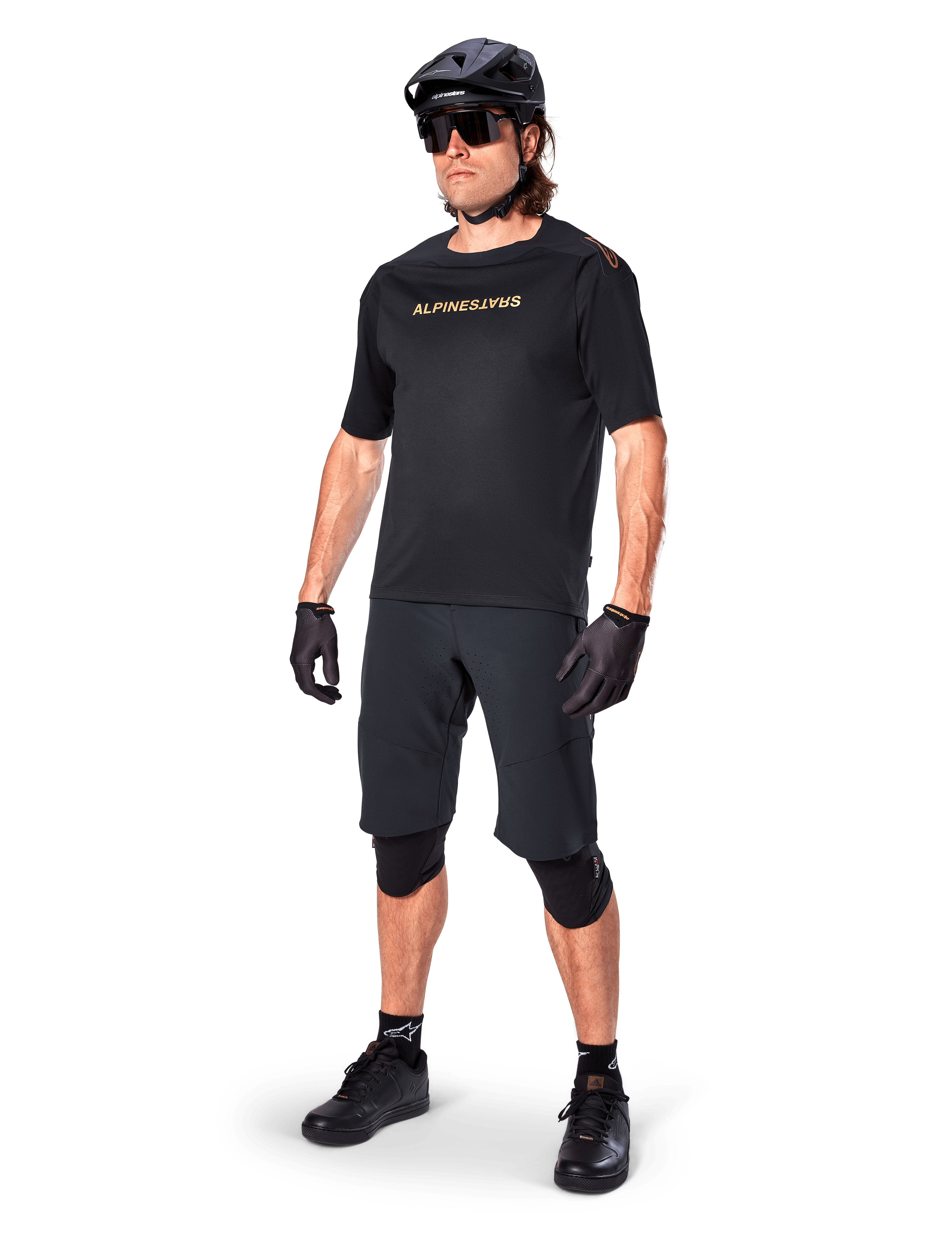 Alpinestars A-Aria Polartec Switch Short Sleeve Jersey, maglia a maniche corte, nero con marchio dorato specchiato sul petto, indossata con pantaloncini da ciclismo neri, casco e ginocchiere, abbigliamento tecnico da MTB per prestazioni fuoristrada
