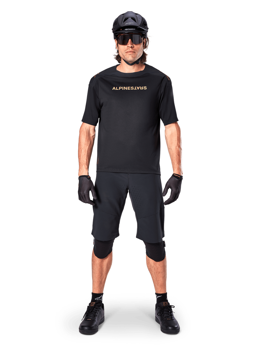 Alpinestars A-Aria Polartec Switch Jersey, maglia MTB a maniche corte, nero con scritte tan, indossata con equipaggiamento da mountain bike nero coordinato comprensivo di casco, pantaloncini e ginocchiere, abbigliamento tecnico da ciclismo fuoristrada