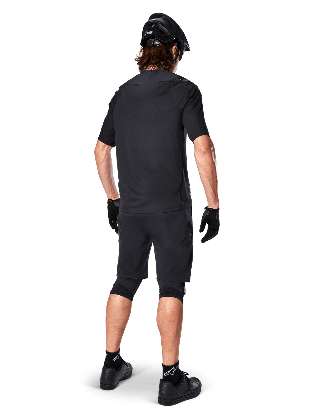 Ciclista di mountain bike visto da dietro con maglia Alpinestars A-Aria Polartec Switch in nero, maglia MTB a maniche corte, abbinata a pantaloncini da mountain bike neri, casco e guanti neri, outfit tecnico coordinato per il trail riding fuoristrada