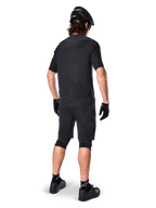 Ciclista di mountain bike visto da dietro con maglia Alpinestars A-Aria Polartec Switch in nero, maglia MTB a maniche corte, abbinata a pantaloncini da mountain bike neri, casco e guanti neri, outfit tecnico coordinato per il trail riding fuoristrada
