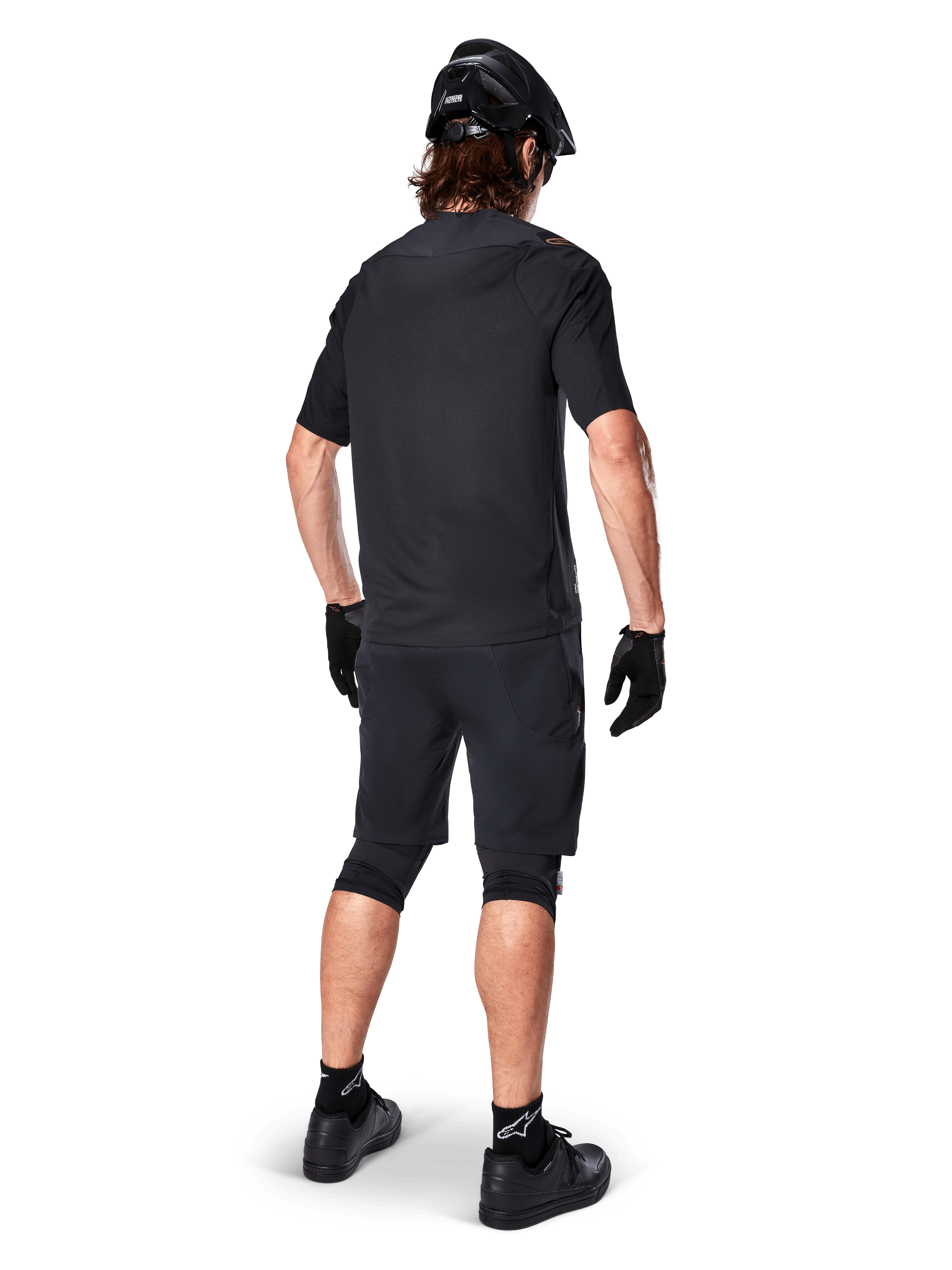 Ciclista di mountain bike visto da dietro con maglia Alpinestars A-Aria Polartec Switch in nero, maglia MTB a maniche corte, abbinata a pantaloncini da mountain bike neri, casco e guanti neri, outfit tecnico coordinato per il trail riding fuoristrada