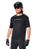 Alpinestars A-Aria Polartec Switch Jersey a maniche corte, maglia da mountain bike nera con logo Alpinestars color oro, indossata con casco nero, occhiali da sole e guanti, abbigliamento tecnico da MTB