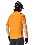 Maglia Alpinestars A-Aria Polartec Switch, maglia da MTB a maniche corte, oro scuro, vista posteriore con vestibilità performante, indossata con casco nero e pantaloncini grigi, abbigliamento da trail riding resistente e traspirante
