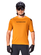 Maglia a maniche corte Alpinestars A-Aria Polartec Switch, oro scuro, ciclista di mountain bike con casco nero e occhiali da sole, abbigliamento tecnico da ciclismo per percorsi MTB