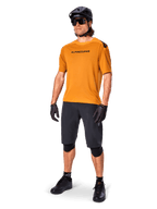 Ciclista che indossa la maglia Alpinestars A-Aria Polartec Switch in Dark Gold, maglia da MTB a maniche corte, abbinata a pantaloncini da ciclismo neri, ginocchiere, casco nero e guanti, abbigliamento professionale da mountain bike per trail riding