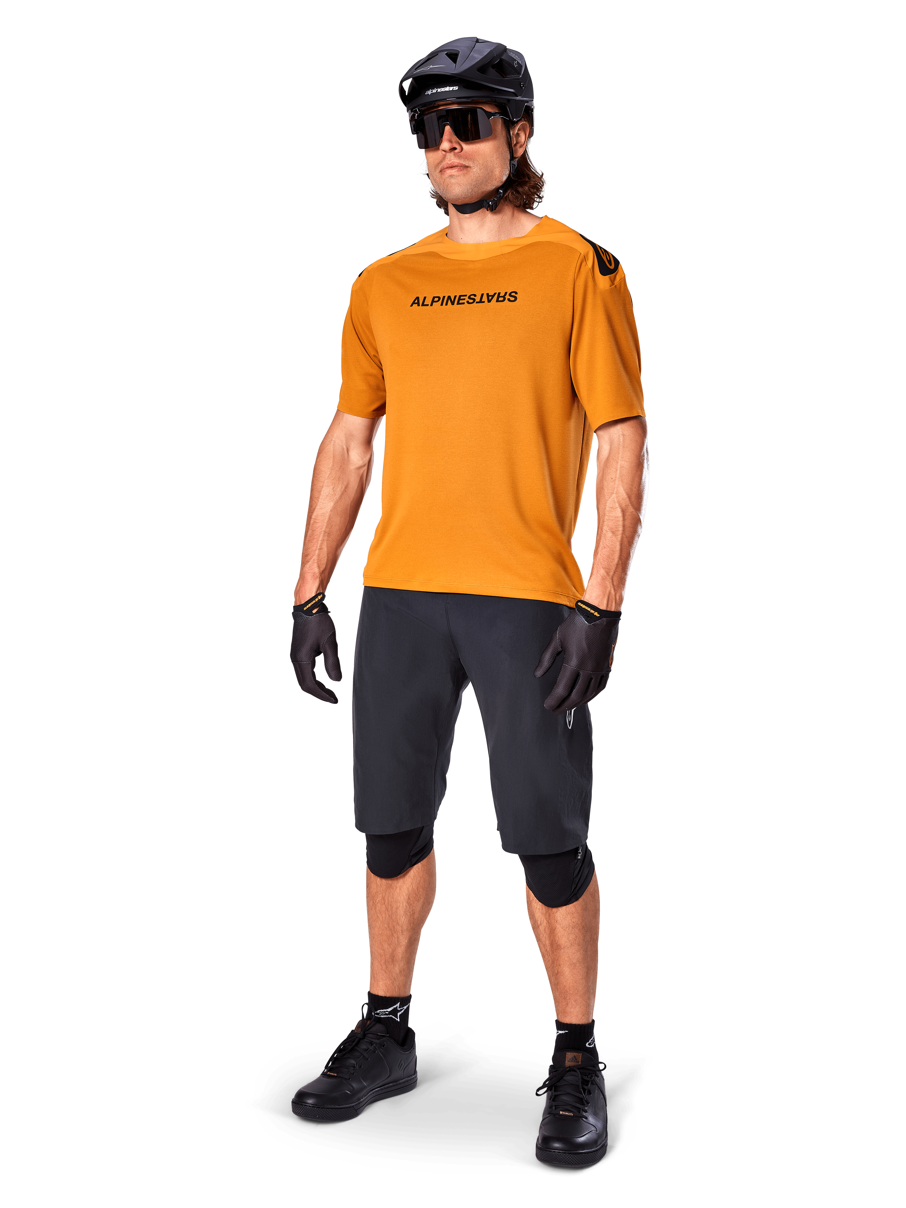 Ciclista che indossa la maglia Alpinestars A-Aria Polartec Switch in Dark Gold, maglia da MTB a maniche corte, abbinata a pantaloncini da ciclismo neri, ginocchiere, casco nero e guanti, abbigliamento professionale da mountain bike per trail riding