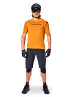 Ciclista uomo che indossa la maglia a maniche corte Alpinestars A-Aria Polartec Switch in Dark Gold, abbinata a pantaloncini da MTB neri, casco, occhiali da sole e guanti, abbigliamento professionale da mountain bike