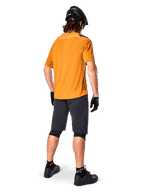 Alpinestars Maglia A-Aria Polartec Switch, maglia MTB a maniche corte, oro scuro, vista posteriore del modello con pantaloncini da mountain bike grigio scuro, casco nero e guanti, abbigliamento tecnico da ciclismo per percorsi fuoristrada