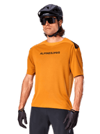 Maglia Alpinestars A-Aria Polartec Switch a maniche corte, color oro scuro con logo nero, indossata da un modello con casco nero, occhiali da sole e guanti da ciclismo, abbigliamento da mountain bike ad alte prestazioni