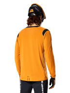 Alpinestars A-Dura Dri Wool Long Sleeve Jersey in Dark Gold, vista posteriore di un ciclista con casco da mountain bike e guanti, con pannelli in rete nera sulle spalle e coda più lunga, abbigliamento tecnico da ciclismo per trail riding
