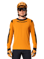 Modello che indossa la maglia a maniche lunghe Alpinestars A-Dura Dri Wool in Dark Gold, coordinata con casco da MTB e guanti oro abbinati, pannelli in rete nera su spalle e maniche, marchio Alpinestars nero sul petto, occhiali da sole e pantaloni scuri, abbigliamento tecnico da mountain bike