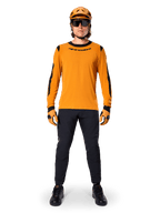 Mountain biker con maglia Alpinestars A-Dura Dri Wool a maniche lunghe, oro scuro con accenti neri su collo e maniche, abbinata a pantaloni neri, casco color oro coordinato, guanti e occhiali da sole neri, abbigliamento professionale da mountain bike