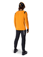 Mountain biker visto da dietro che indossa la maglia Alpinestars A-Dura Dri Wool a maniche lunghe in Dark Gold, pantaloni tecnici neri con logo bianco e un casco coordinato nero e arancione, abbigliamento da mountain bike ad alte prestazioni.
