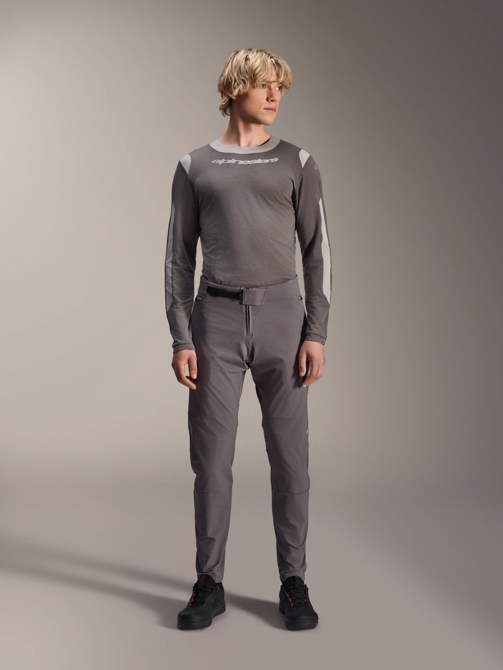 Alpinestars A-Dura Elite Pants, pantaloni da mountain bike grigio scuro, vista a figura intera, indossati con una maglia tecnica coordinata grigia a maniche lunghe con il logo Alpinestars, scarpe nere con dettagli rossi, completo da ciclismo sportivo