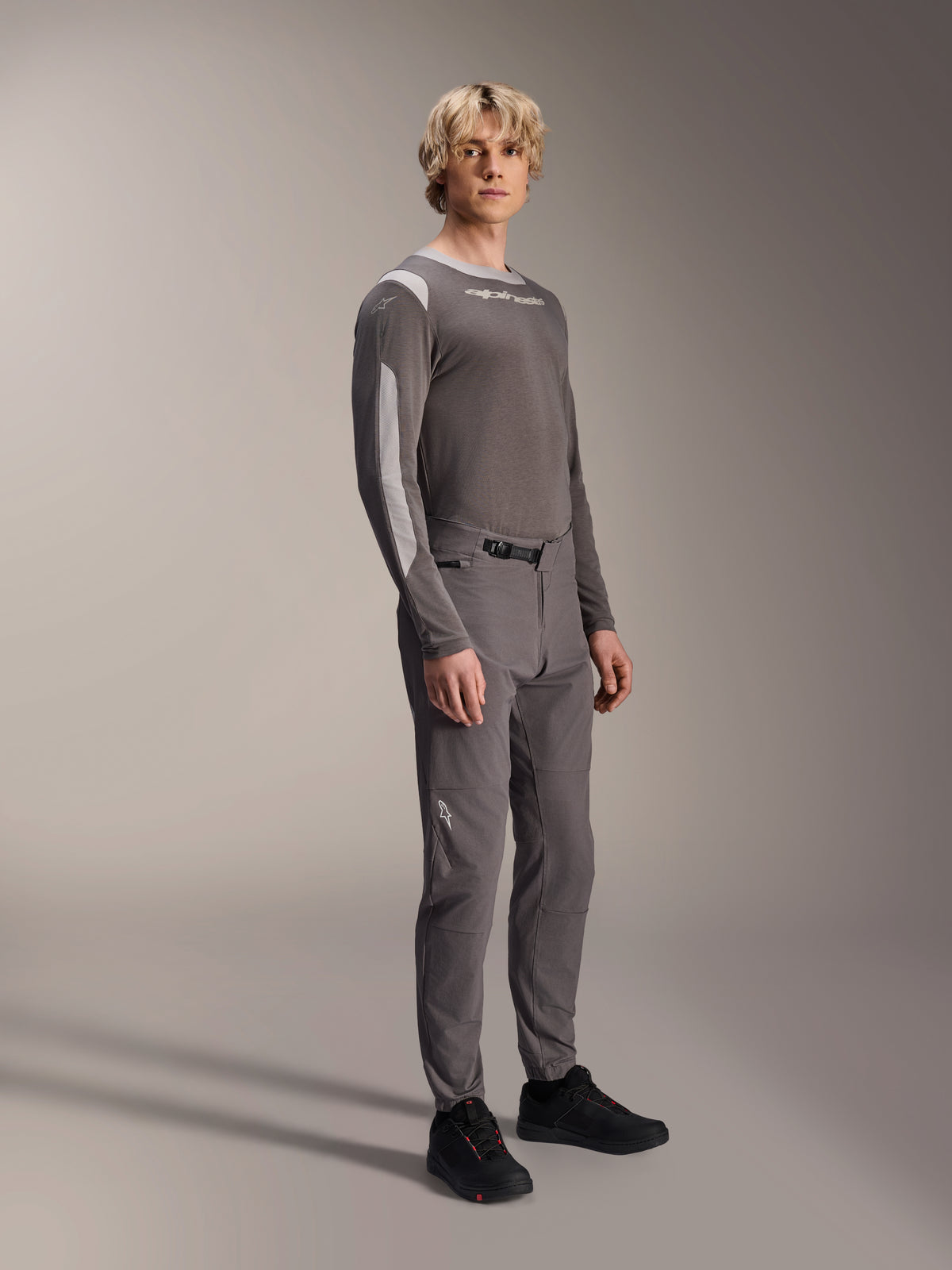 Alpinestars A-Dura Elite Pants, pantaloni da mountain bike, grigio scuro, indossati con maglia coordinata a maniche lunghe e scarpe nere, dotati di chiusura anteriore a cricchetto e tessuto Cordura resistente per trail ed enduro