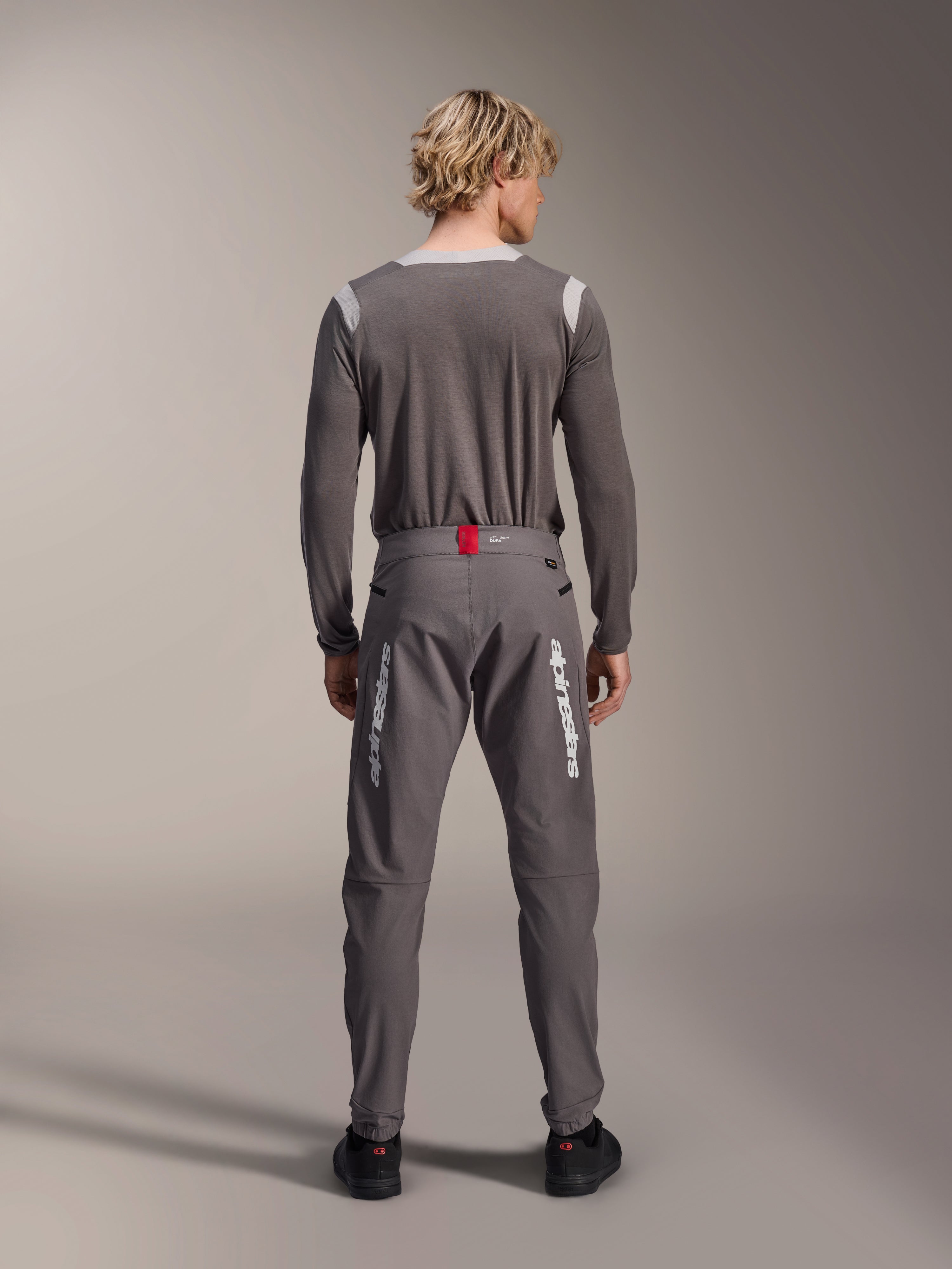 Modello visto da dietro che indossa pantaloni Alpinestars A-Dura Elite grigio scuro e una maglia grigia coordinata a maniche lunghe, con loghi Alpinestars bianchi sulle gambe, dettaglio rosso in vita e scarpe nere, abbigliamento professionale da MTB.
