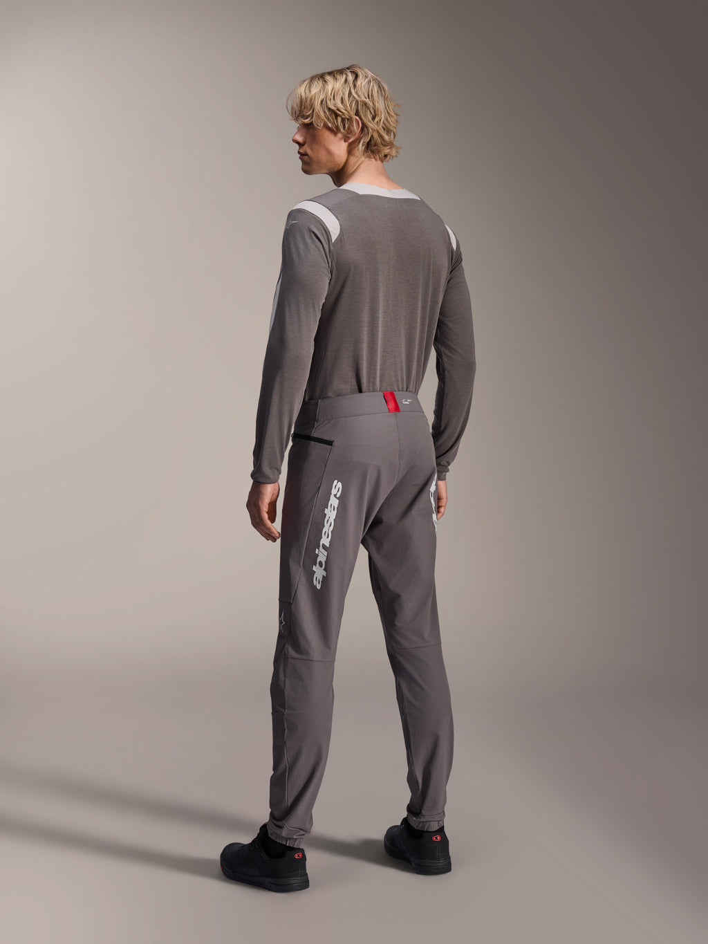 Modello visto da dietro con abbigliamento Alpinestars MTB, maglia a maniche lunghe A-Dura Dri Wool grigia, pantaloni A-Dura Elite grigio scuro con logo verticale bianco e dettaglio rosso in vita, completo coordinato da mountain bike