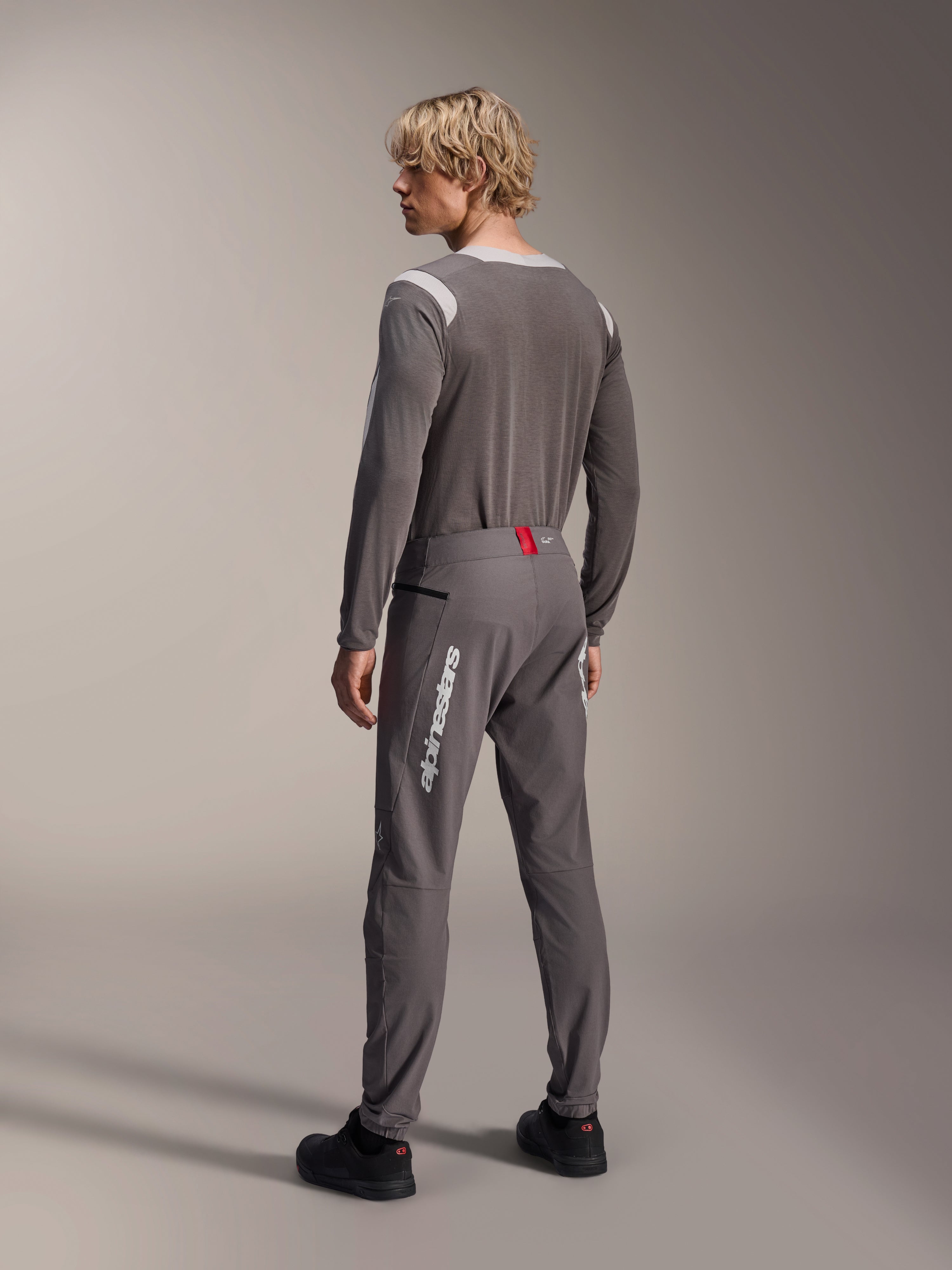 Modello visto da dietro con abbigliamento Alpinestars MTB, maglia a maniche lunghe A-Dura Dri Wool grigia, pantaloni A-Dura Elite grigio scuro con logo verticale bianco e dettaglio rosso in vita, completo coordinato da mountain bike