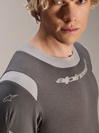 Alpinestars Maglia A-Dura Dri Wool a maniche lunghe, maglia da mountain bike grigio scuro, primo piano di un modello maschile che mostra il colletto elasticizzato e i pannelli in rete sulle spalle, con logo Alpinestars sul petto e sulla manica