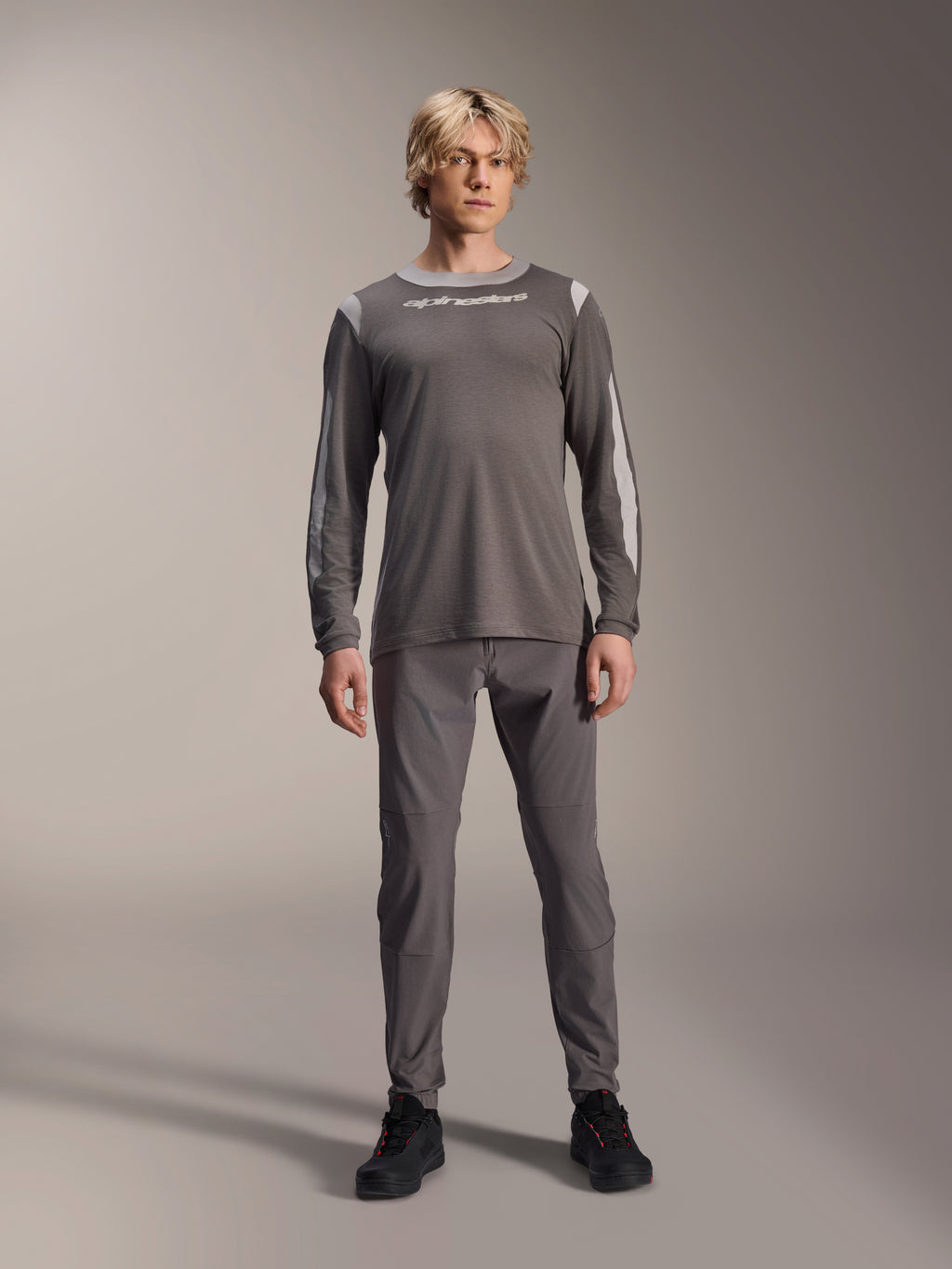 Maglia a maniche lunghe Alpinestars A-Dura Dri Wool, grigio scuro, indossata da un modello con pantaloni grigi coordinati e scarpe nere, logo Alpinestars bianco sul petto, pannelli in rete grigio chiaro su spalle e maniche, abbigliamento tecnico per mountain bike