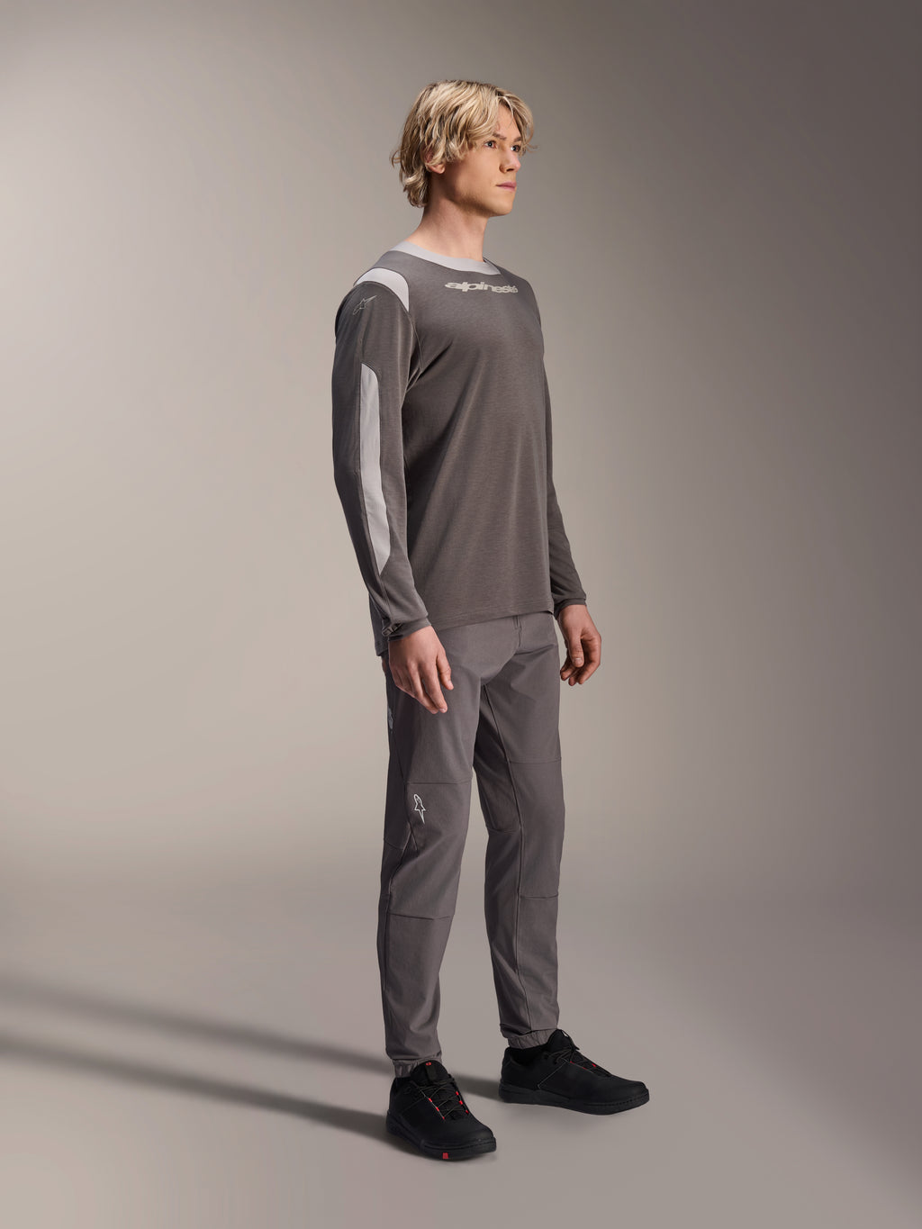 Modello con maglia Alpinestars A-Dura Dri Wool a maniche lunghe in grigio scuro, pantaloni grigi coordinati e scarpe nere, con pannelli in rete grigio chiaro su spalle e maniche, abbigliamento tecnico da MTB per il ciclismo fuoristrada