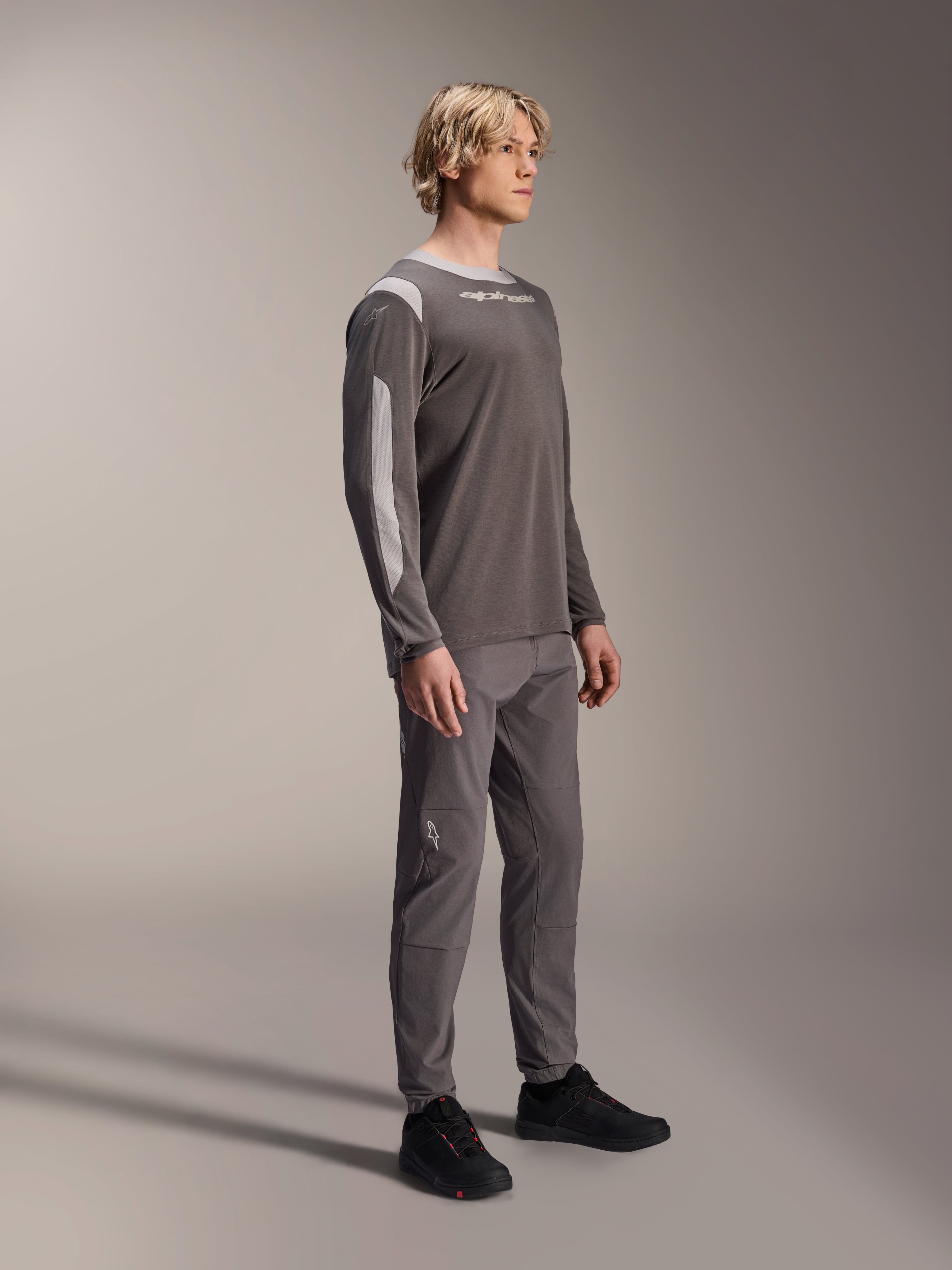 Modello con maglia Alpinestars A-Dura Dri Wool a maniche lunghe in grigio scuro, pantaloni grigi coordinati e scarpe nere, con pannelli in rete grigio chiaro su spalle e maniche, abbigliamento tecnico da MTB per il ciclismo fuoristrada