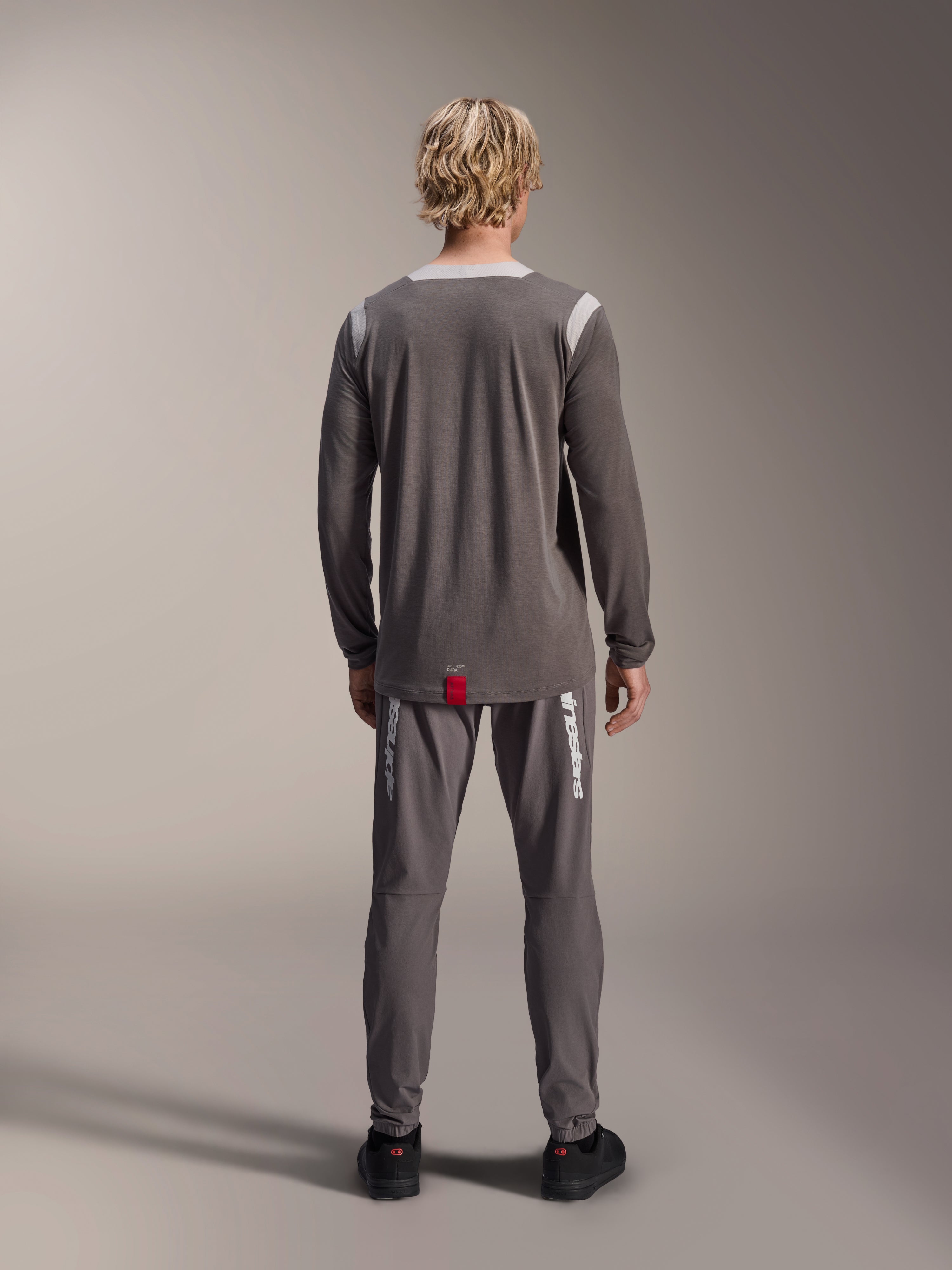 Maglia Alpinestars A-Dura Dri Wool a maniche lunghe, grigio scuro, vista posteriore su modello, con pannelli in rete grigio chiaro su spalle e colletto, bordo posteriore più lungo con piccola etichetta rossa del brand, indossata con pantaloni da ciclismo grigi coordinati
