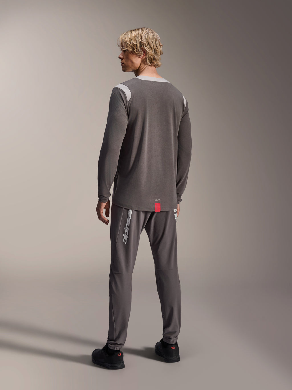 Modello che indossa la maglia a maniche lunghe Alpinestars A-Dura Dri Wool, grigio scuro con pannelli in rete grigio chiaro sulle spalle, abbinata a pantaloni da ciclismo coordinati, vista posteriore con orlo allungato, abbigliamento mountain bike