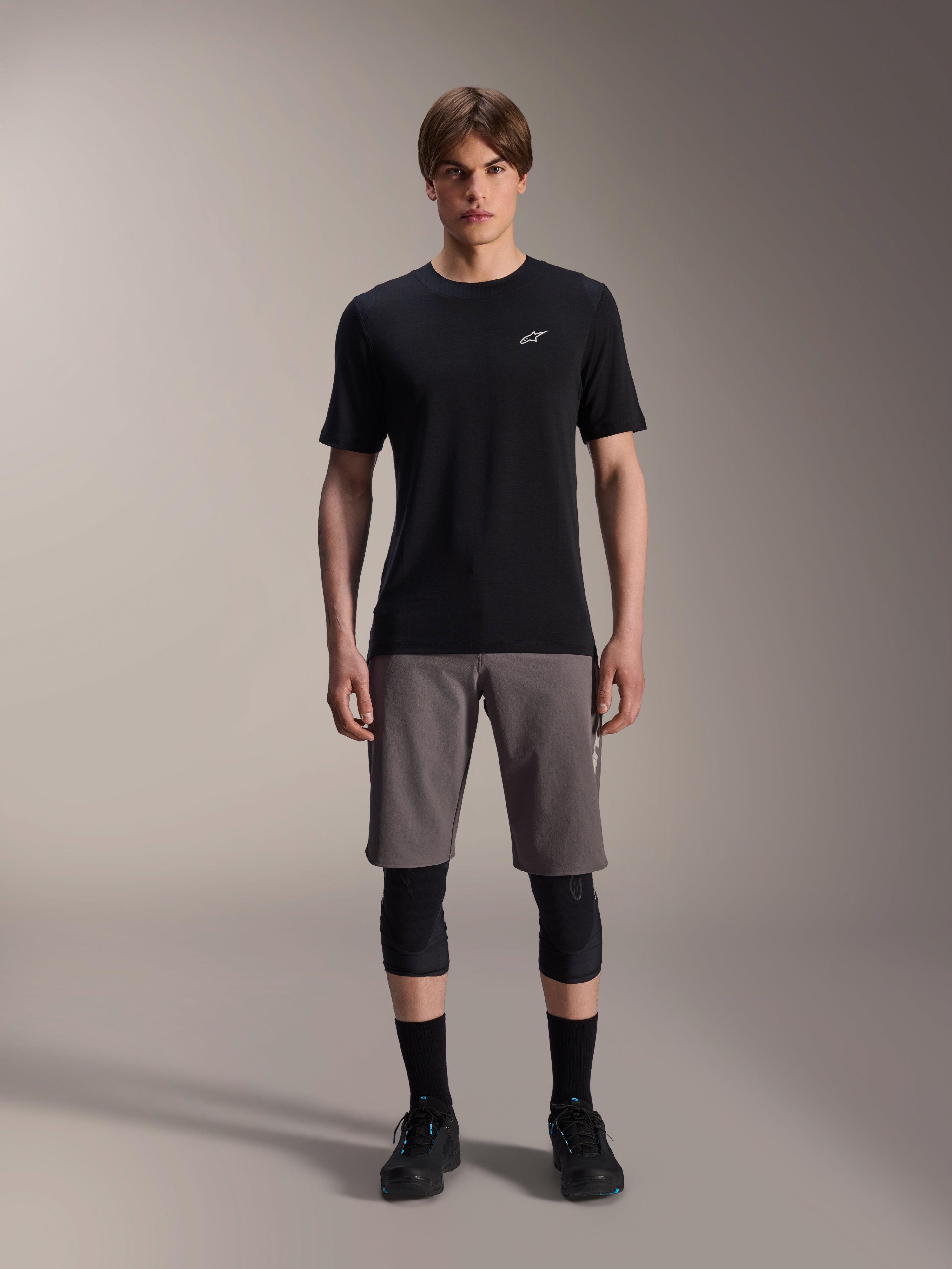 Modello con pantaloncini Alpinestars A-Dura Elite in grigio scuro e maglia a maniche corte A-Dura Dri-Wool in nero, abbinati a ginocchiere nere e scarpe da ciclismo, abbigliamento tecnico da mountain bike per prestazioni off-road