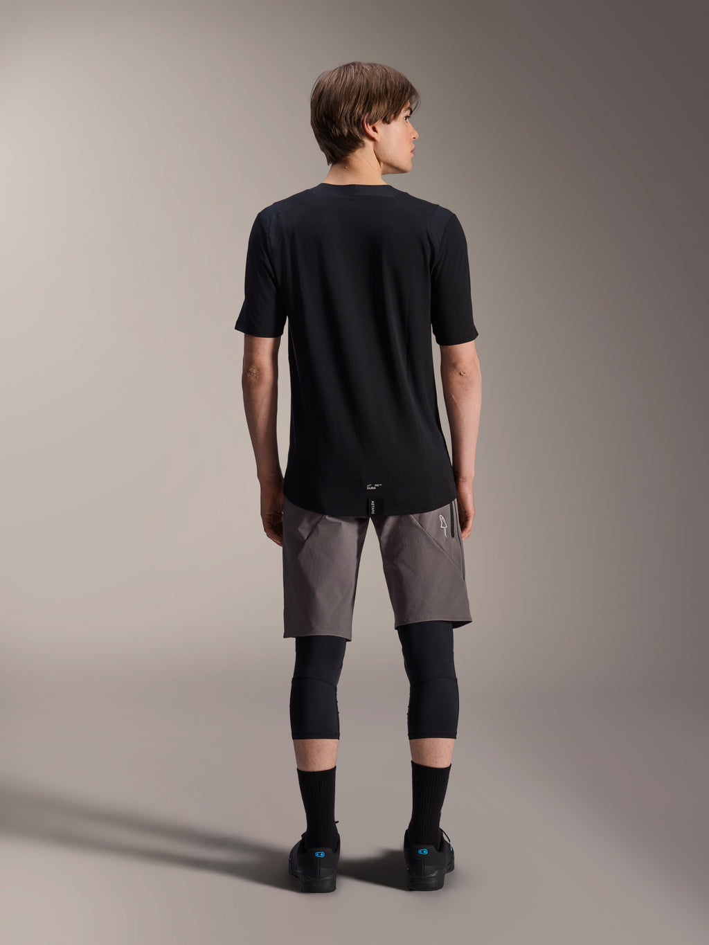 Alpinestars A-Dura Elite Shorts, pantaloncini da mountain bike grigio scuro, vista posteriore, indossati con maglia nera a maniche corte, ginocchiere e calze nere, abbigliamento tecnico da ciclismo per il fuoristrada