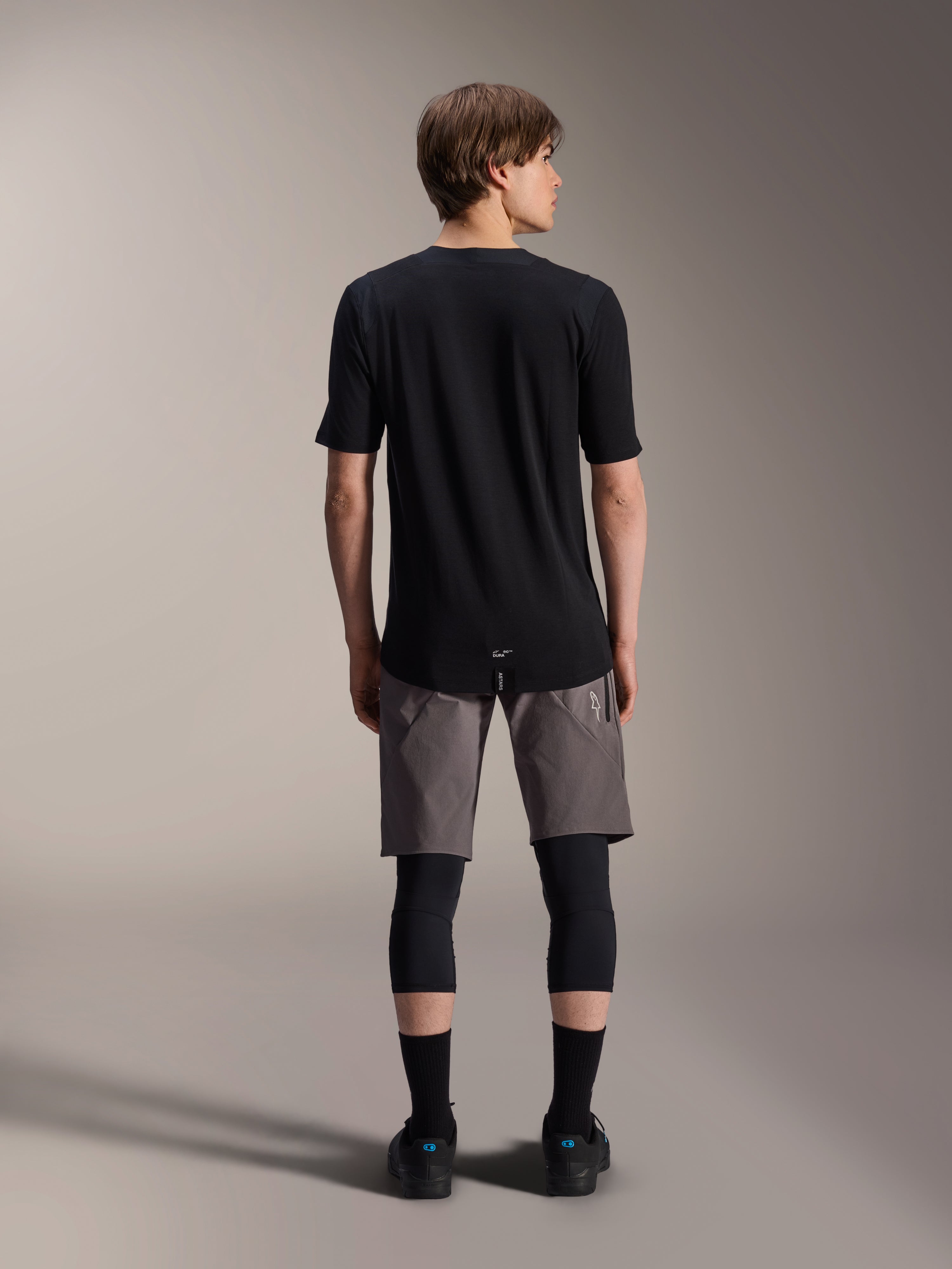 Alpinestars A-Dura Elite Shorts, pantaloncini da mountain bike grigio scuro, vista posteriore, indossati con maglia nera a maniche corte, ginocchiere e calze nere, abbigliamento tecnico da ciclismo per il fuoristrada