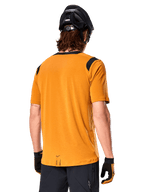 Alpinestars Maglia A-Dura Dri Wool a maniche corte, Dark Gold, vista posteriore di un uomo che indossa la maglia con pannelli in rete neri sulle spalle, abbinata a casco e guanti da MTB neri, abbigliamento da mountain bike con vestibilità regolare