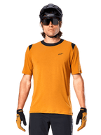 Maglia a maniche corte Alpinestars A-Dura Dri Wool, Oro Scuro, indossata con casco MTB nero, occhiali da sole e guanti, con colletto e pannelli sulle spalle neri, in misto lana traspirante drirelease per mountain bike